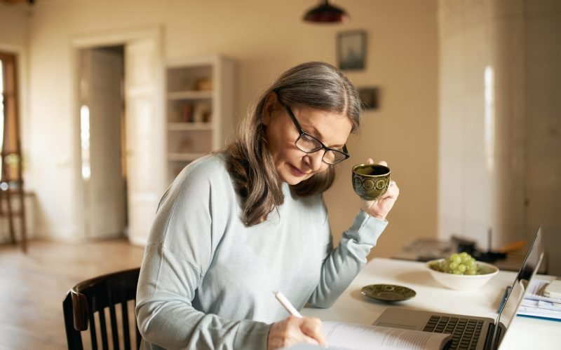 Concentrated middle aged female teacher in glasses sitting at desk drinking coffee, handwriting in copybook, making lesson plan, using portable computer, teaching online from home. Age and technology