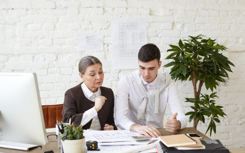 Picture of beautiful experienced elderly woman chief architect helping her skillful young man trainee with architectural project, revising his drawings and pointing at mistakes and drawbacks
