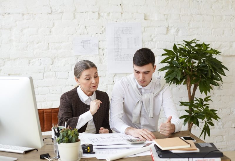 Picture of beautiful experienced elderly woman chief architect helping her skillful young man trainee with architectural project, revising his drawings and pointing at mistakes and drawbacks